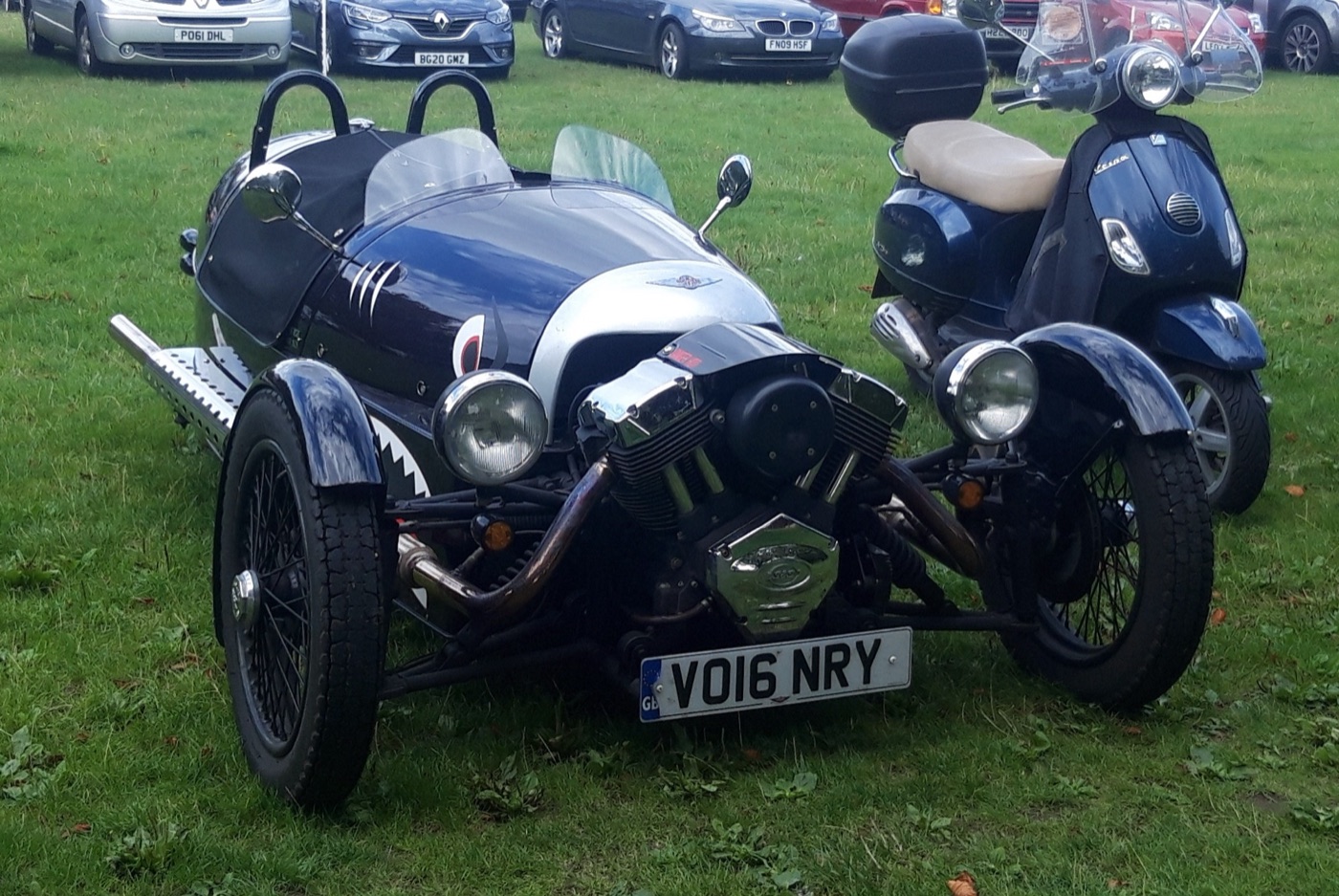 Dark blue Morgan 3 Wheeler with shark-mouth graphics on its nose, chrome V-twin engine and wire wheels, parked on grass next to a scooter