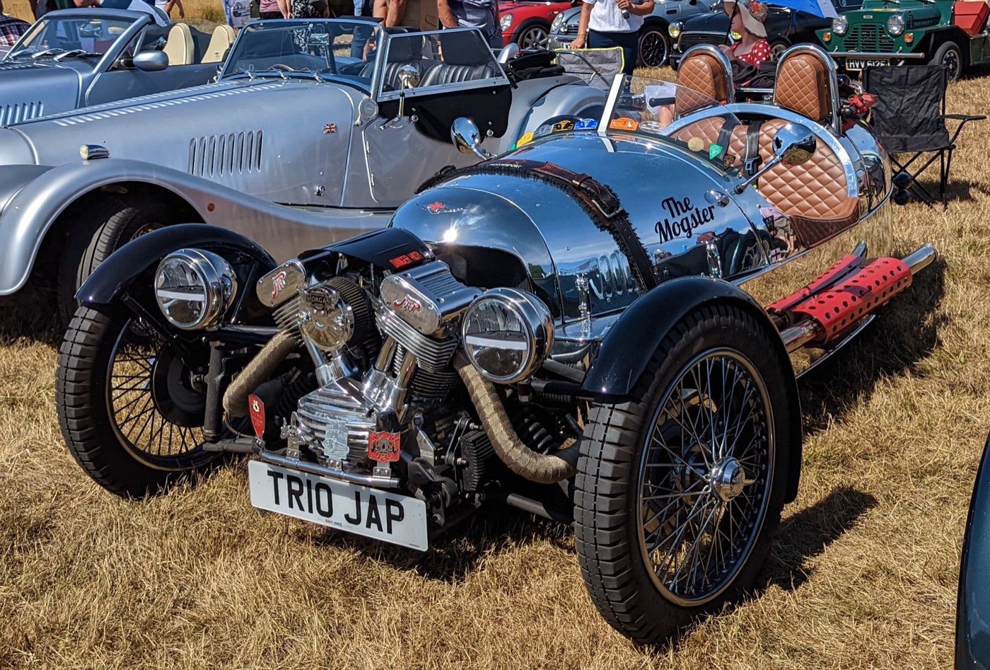 Morgan 3 Wheeler with a polished aluminium body, exposed chrome V-twin engine, wire wheels and quilted tan seats, parked on grass at a car show