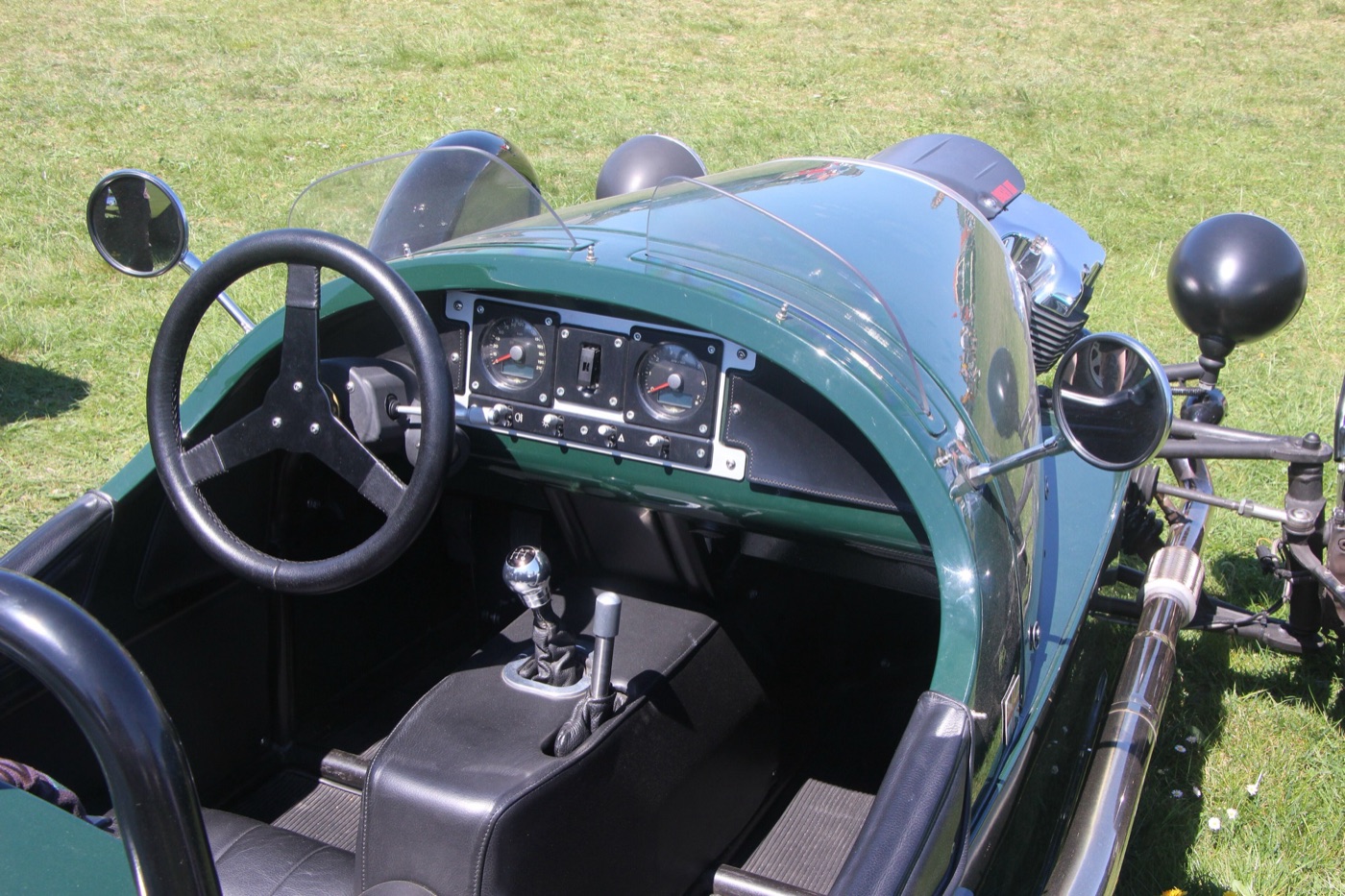 Cockpit of a green Morgan 3 Wheeler: three-spoke steering wheel, two dials and toggle switches on a dark panel, gear lever, and the V-twin beyond the scuttle
