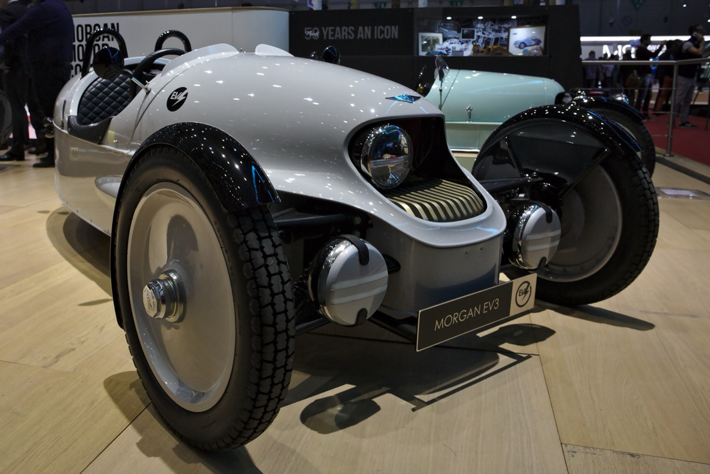 Silver Morgan EV3 electric three-wheeler with brass-finned nose, single round headlamp and two front wheels on a show stand