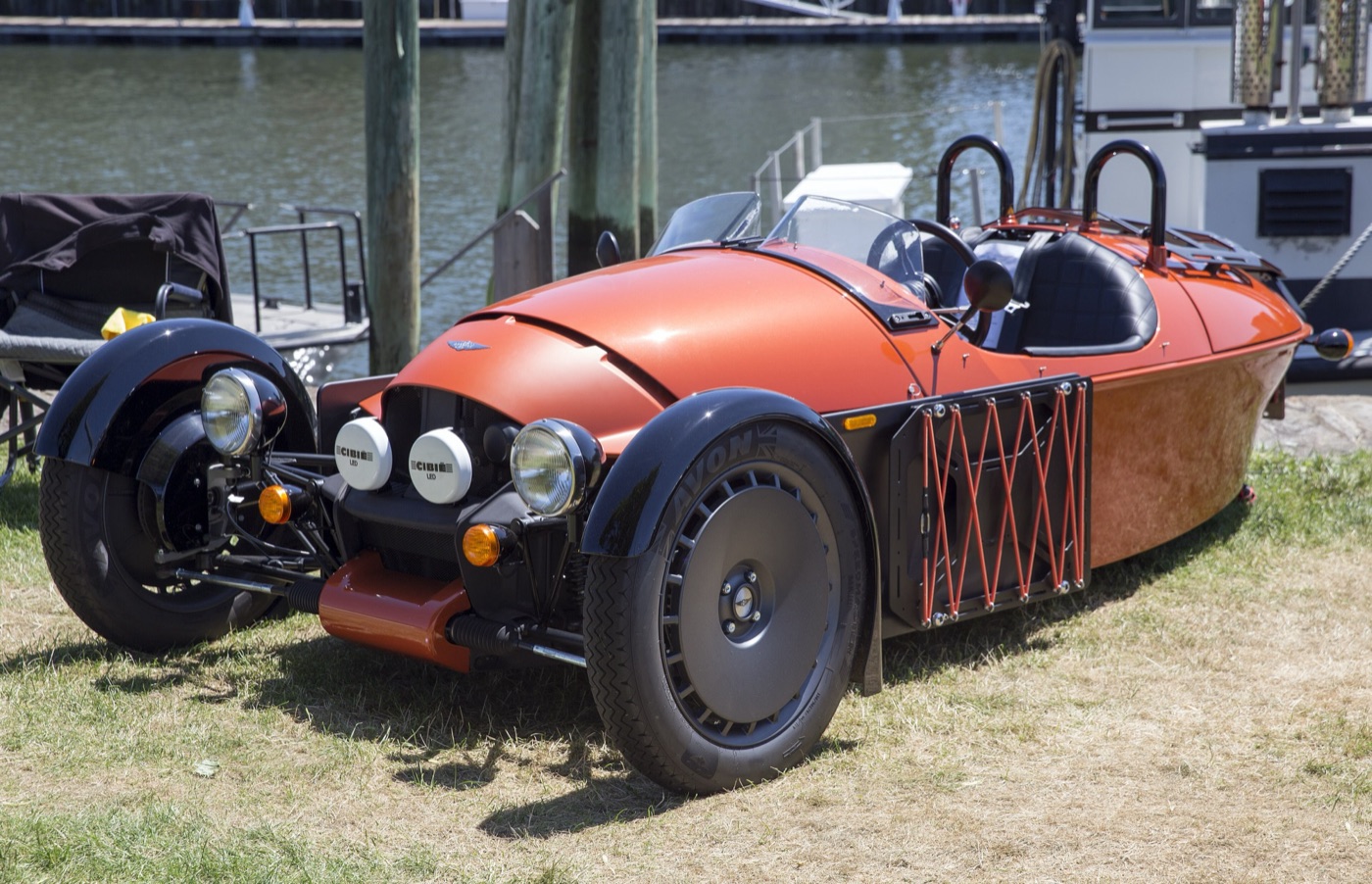 Coral orange Morgan Super 3 with black front wings, painted nose, side luggage blade and two roll hoops, parked by a harbour