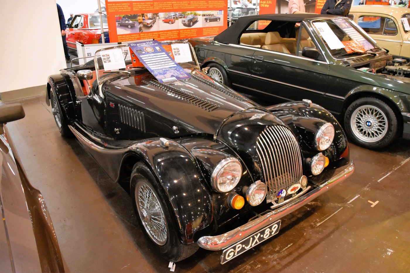 Black 1975 Morgan Plus 8 roadster with wire wheels, a louvred bonnet and a chrome grille on display at an indoor show