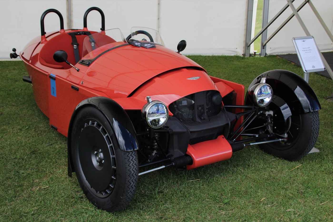 Orange Morgan Super 3 three-wheeler with black wheels and twin roll hoops on grass inside a show marquee