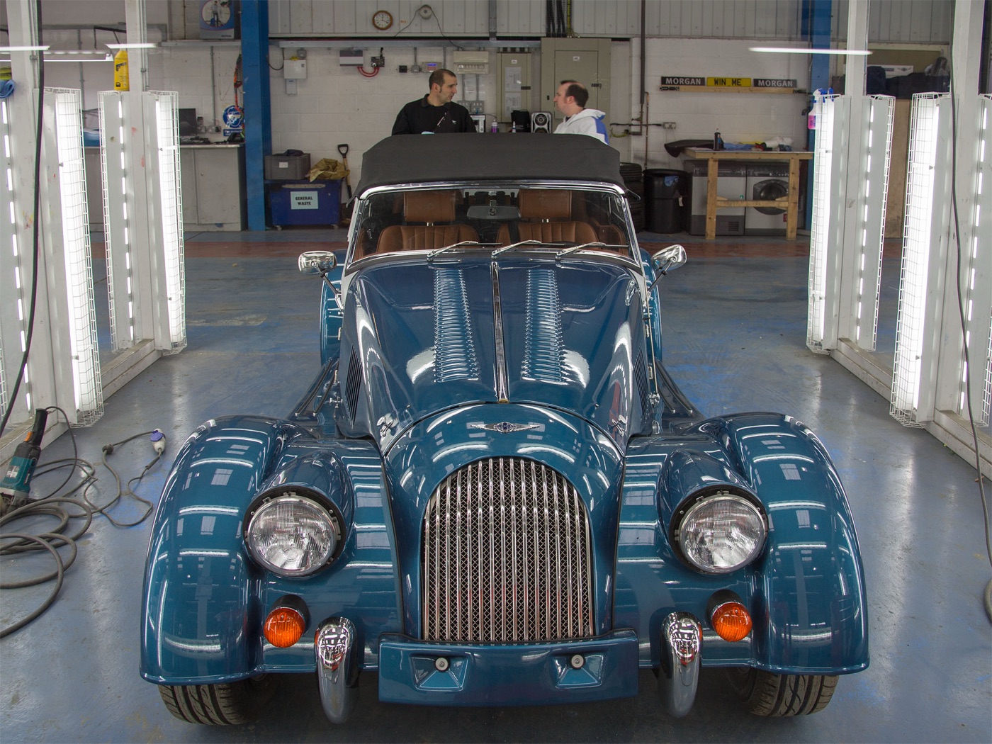 Blue traditional Morgan roadster with its hood up and chrome grille under the inspection lights at the end of the line in the Malvern factory