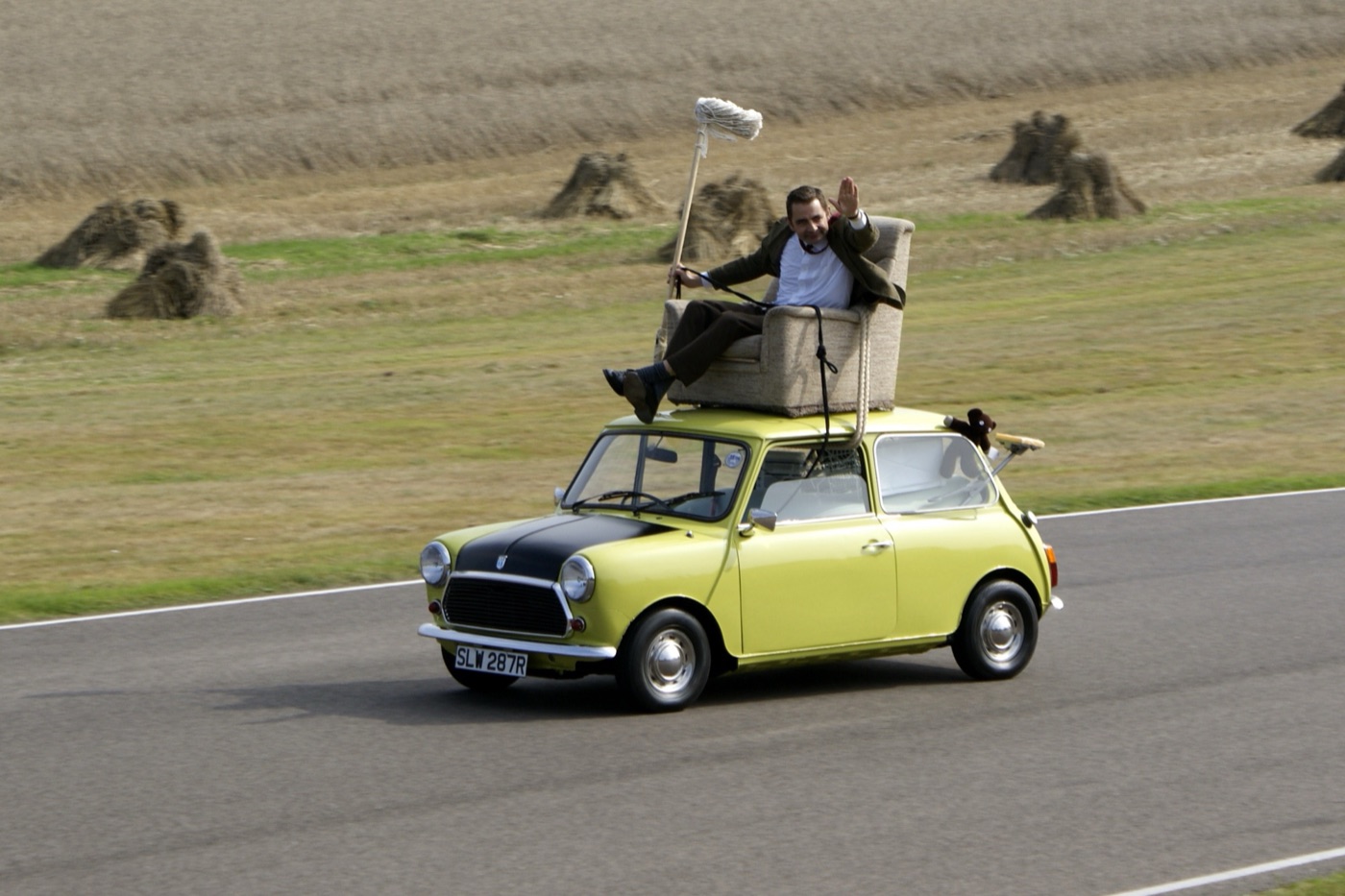 Mr. Bean waving from an armchair strapped to the roof of a green Mini with a black bonnet and the plate SLW 287R, holding a mop, on the Goodwood circuit