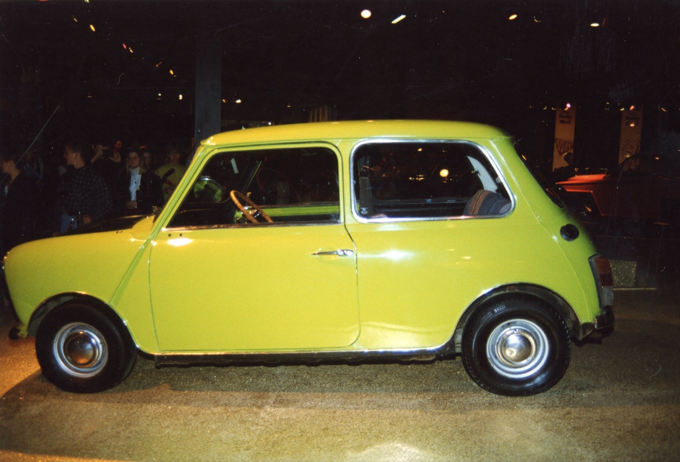 Side view of a Citron Green Mini 1000 with chrome hubcaps and a striped seat cover, on a stand at a busy indoor car show