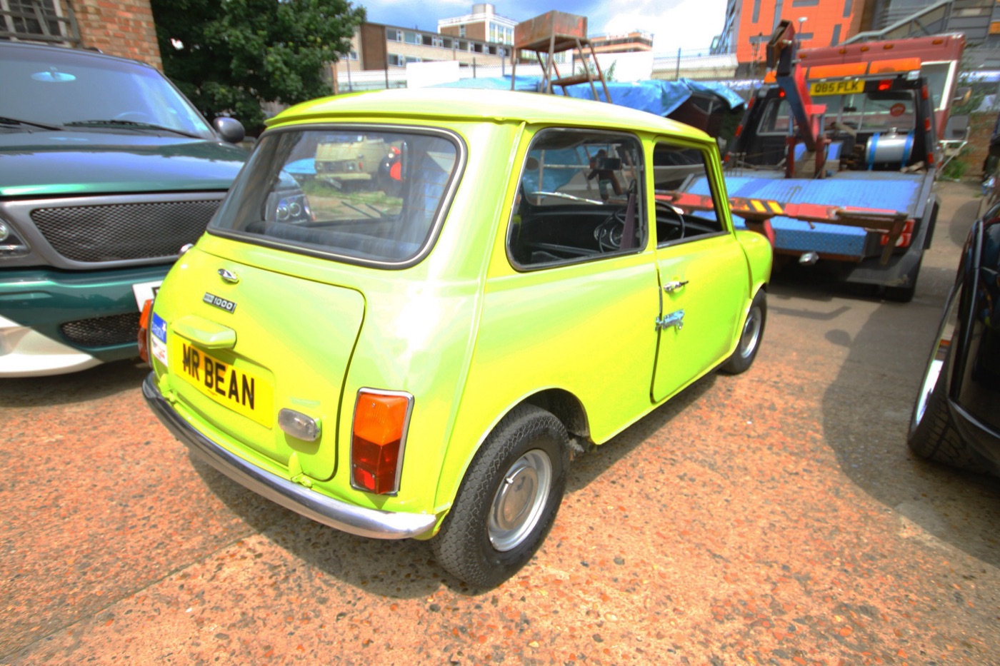 Rear three-quarter view of a lime green Mini 1000 replica with the number plate MR BEAN, parked in a yard beside a recovery truck