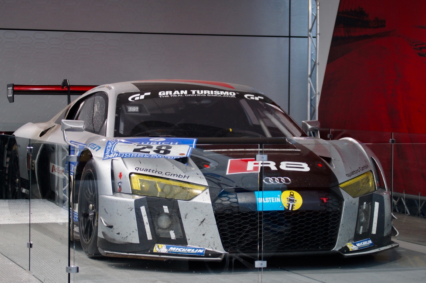Silver and black Audi R8 LMS number 28, still dirty from racing, on display behind glass panels