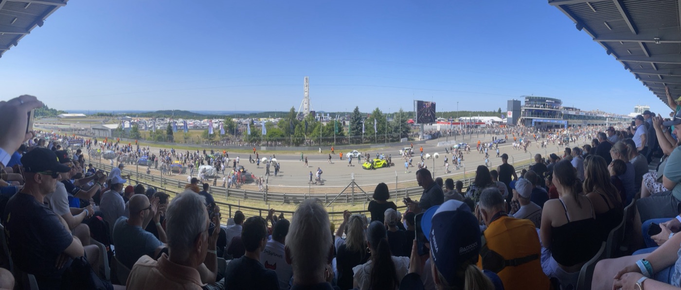 Wide panorama from a covered grandstand of spectators watching the grid walk on the Grand Prix straight, with cars on the grid, a Ferris wheel and the pit building
