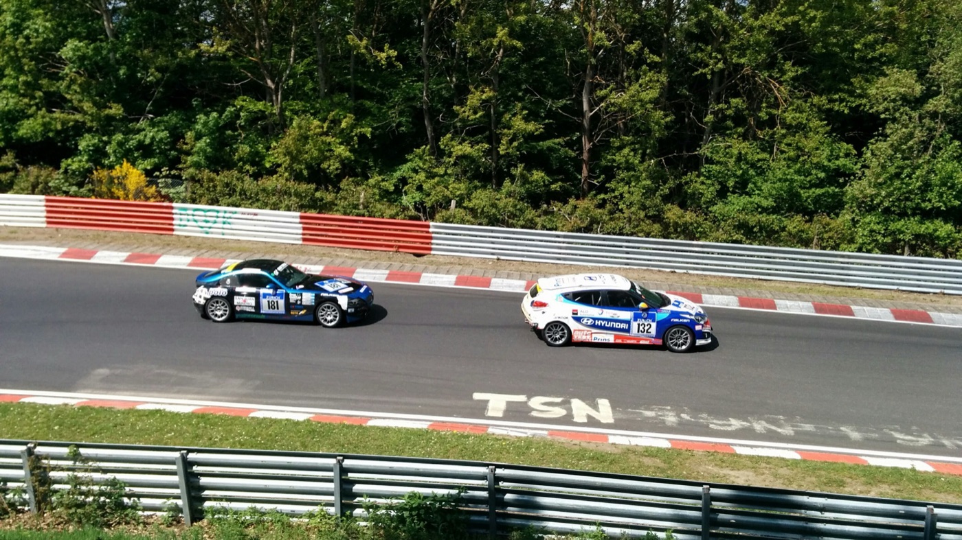 A white Hyundai Veloster number 132 running alongside a dark blue BMW Z4 coupé number 181 between red and white kerbs, with woods behind the barrier