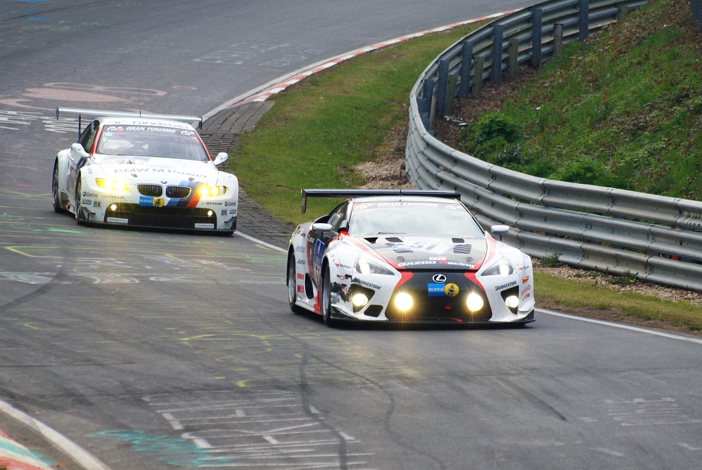 White, red and black Lexus LFA with headlights on, followed by a white BMW M3 race car with BMW Motorsport stripes, on a damp track