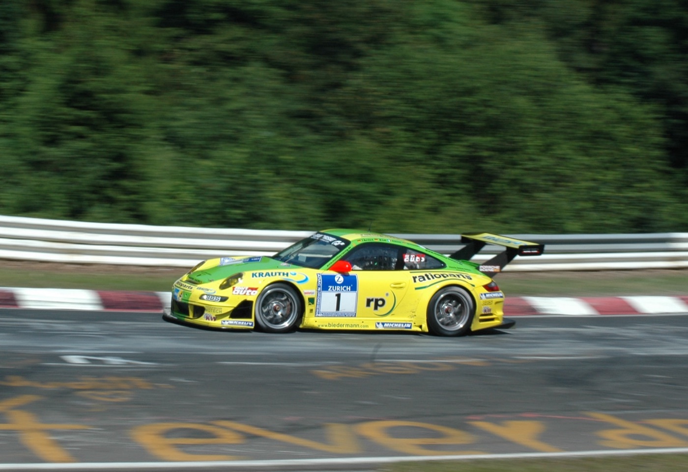 Yellow and green Manthey Racing Porsche 911 GT3 RSR number 1 at speed past the barriers, trees blurred behind