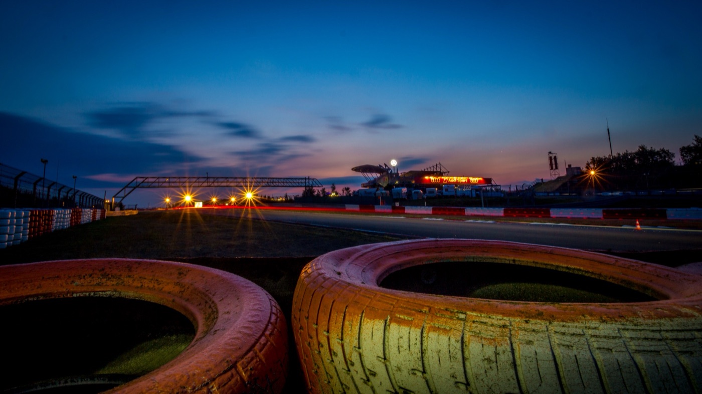 The Grand Prix circuit before dawn, with tyre walls in the foreground, a footbridge over the track and the lit grandstands and Audi Sport sign under a deep blue sky