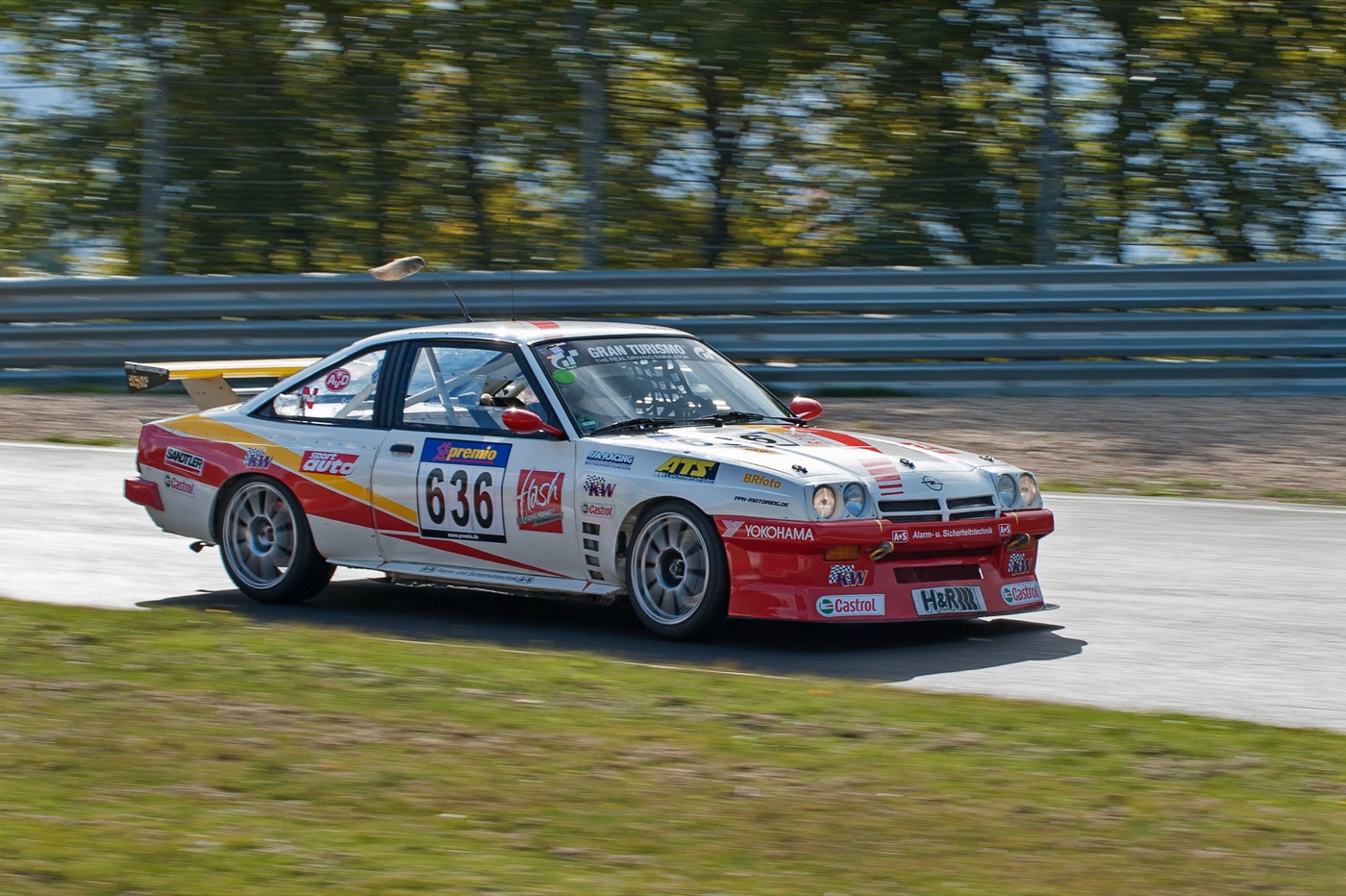 White, red and yellow Opel Manta race car number 636 with a foxtail hanging from its aerial, cornering at speed