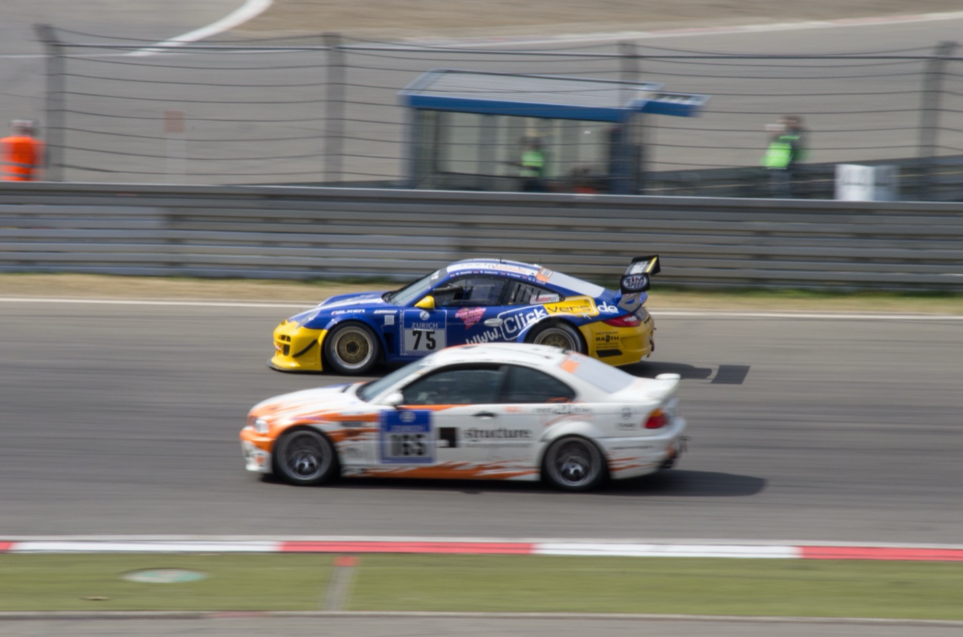A blue and yellow Porsche 911 race car, number 75, and a white and orange BMW M3 side by side at speed on the Grand Prix circuit