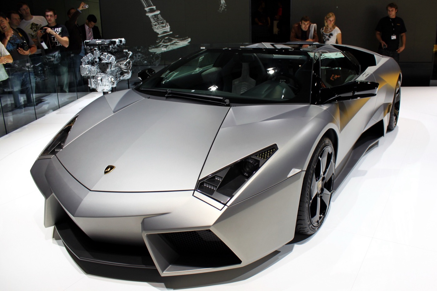 Matt grey Lamborghini Reventón Roadster with its roof open, seen from the front three-quarter on a white stand, with a V12 engine displayed behind it