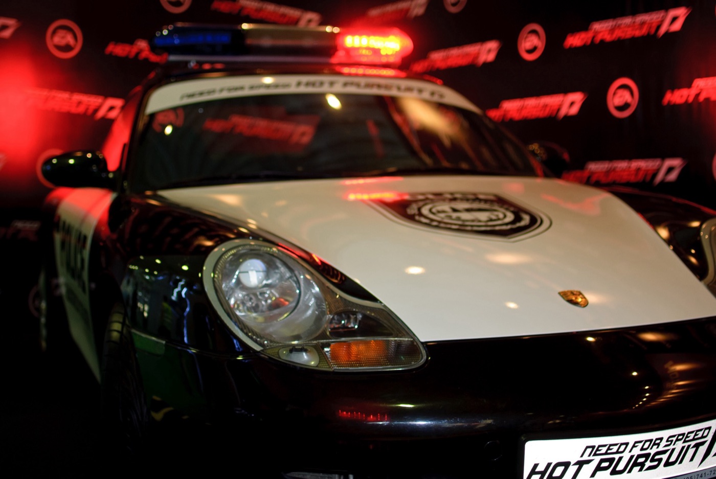 A Porsche painted white and black as a Seacrest County police car, with a light bar on the roof and the game logo on its number plate, on a show stand