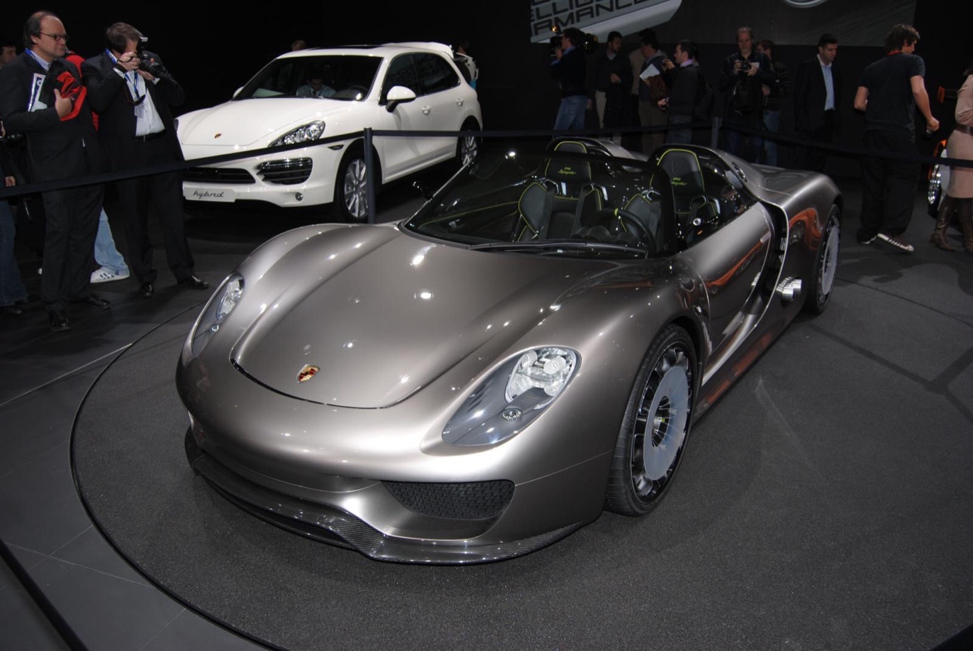 Silver Porsche 918 Spyder concept roadster on a turntable, with photographers and a white Porsche Cayenne behind it