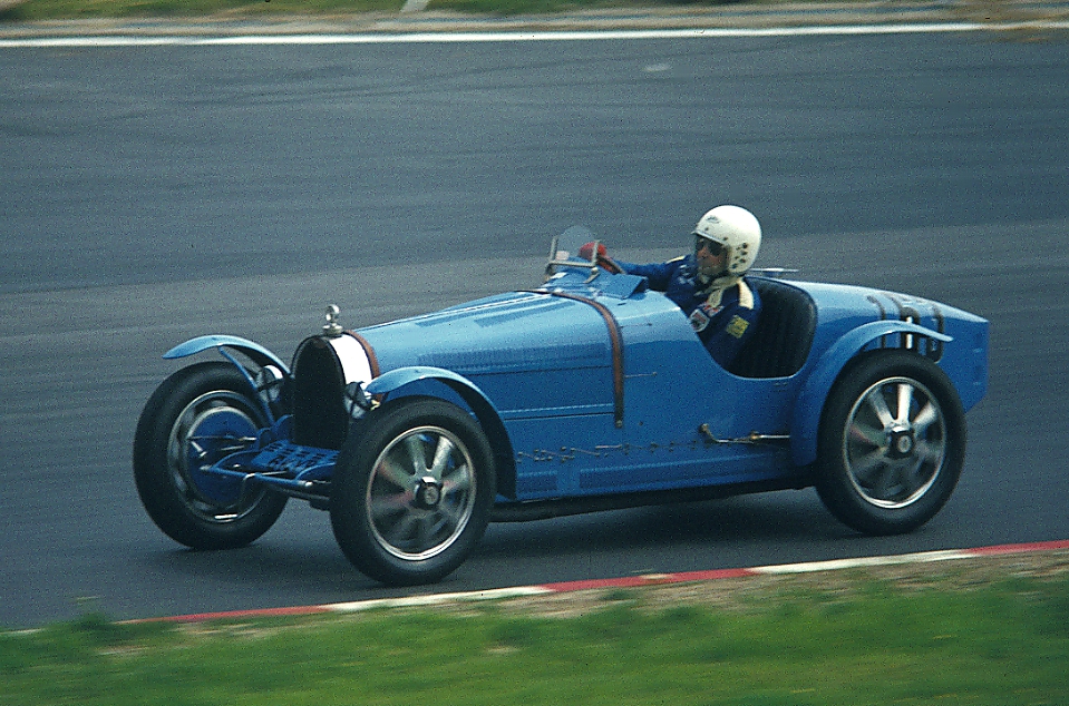 Pale blue Bugatti Type 35B two-seater with alloy wheels being driven along a circuit by a driver in a white helmet