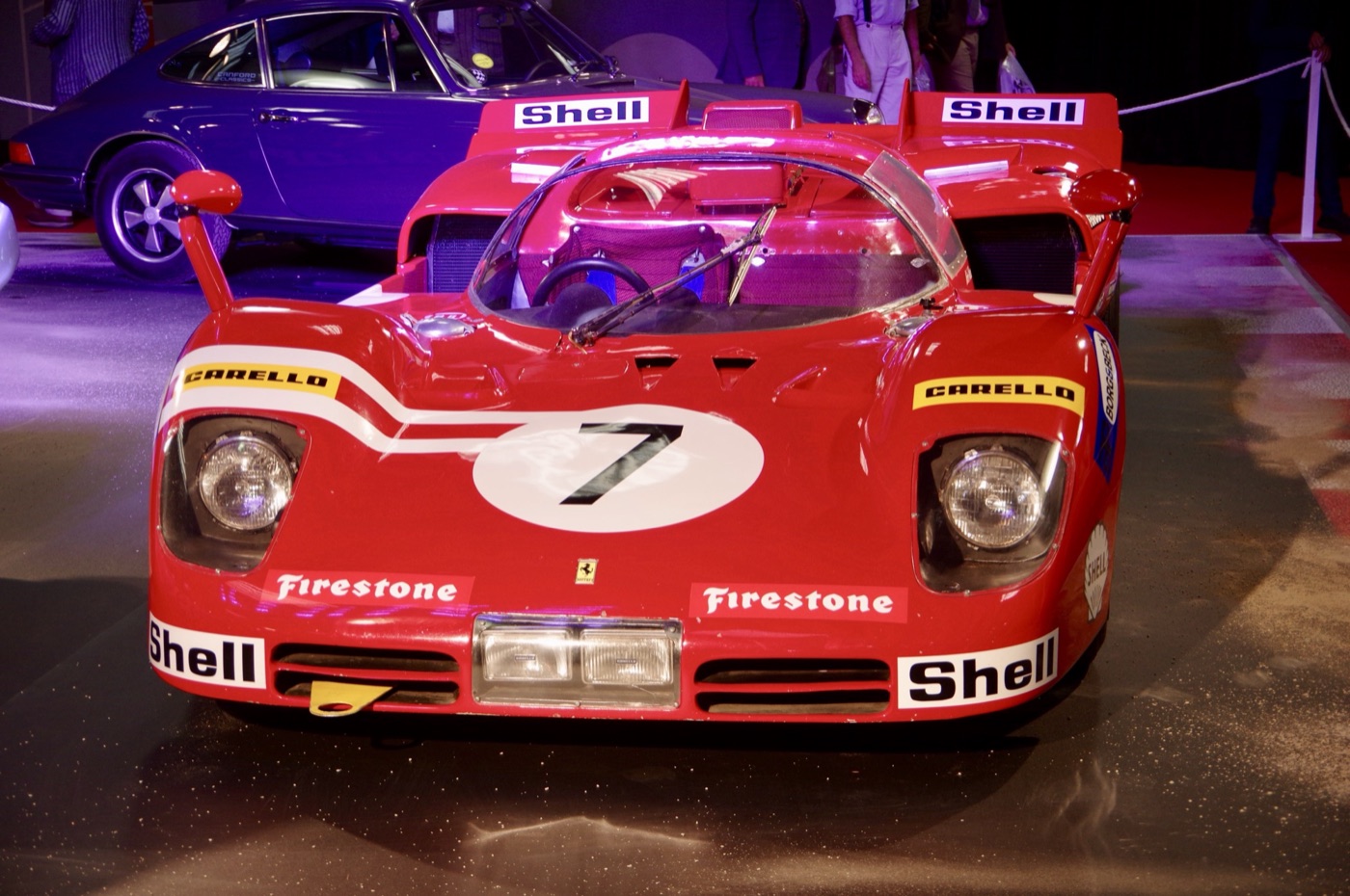 Front view of a red Ferrari 512 S endurance racer with Shell, Firestone and Carello decals and the number 7 on its nose, on display beside a Porsche 911