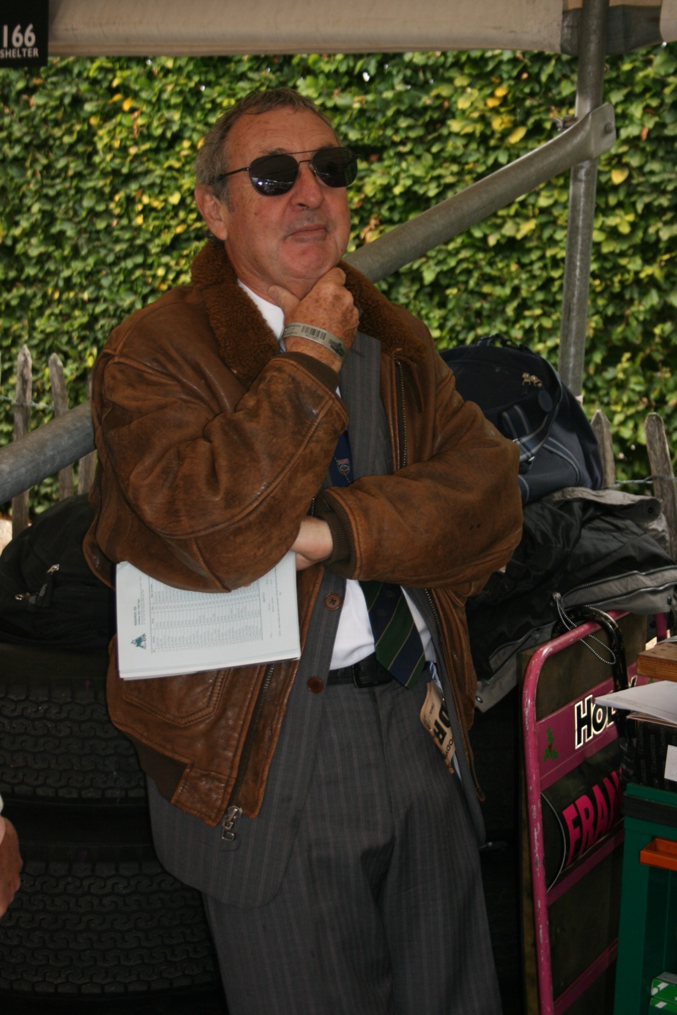 Nick Mason in sunglasses, a brown leather flying jacket and a striped tie, holding a results sheet under his arm beside a paddock shelter
