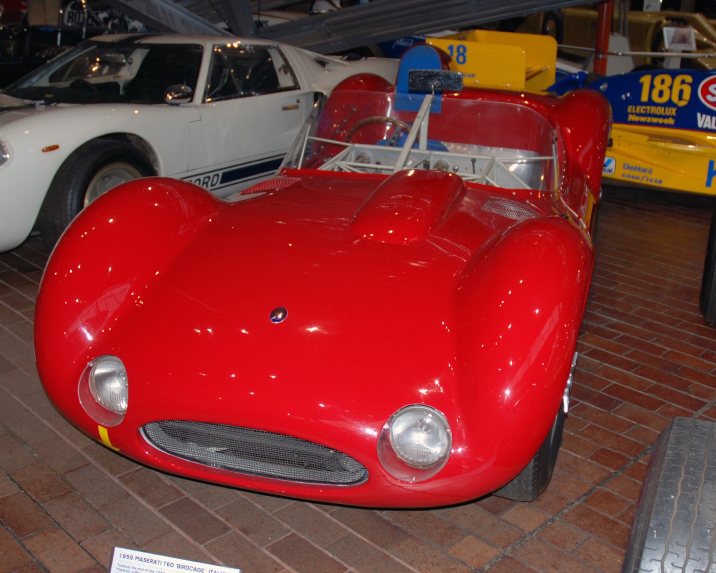 Red Maserati Tipo 60 Birdcage sports-racing car with its tubular frame visible through the windscreen, parked on a brick floor among other racing cars