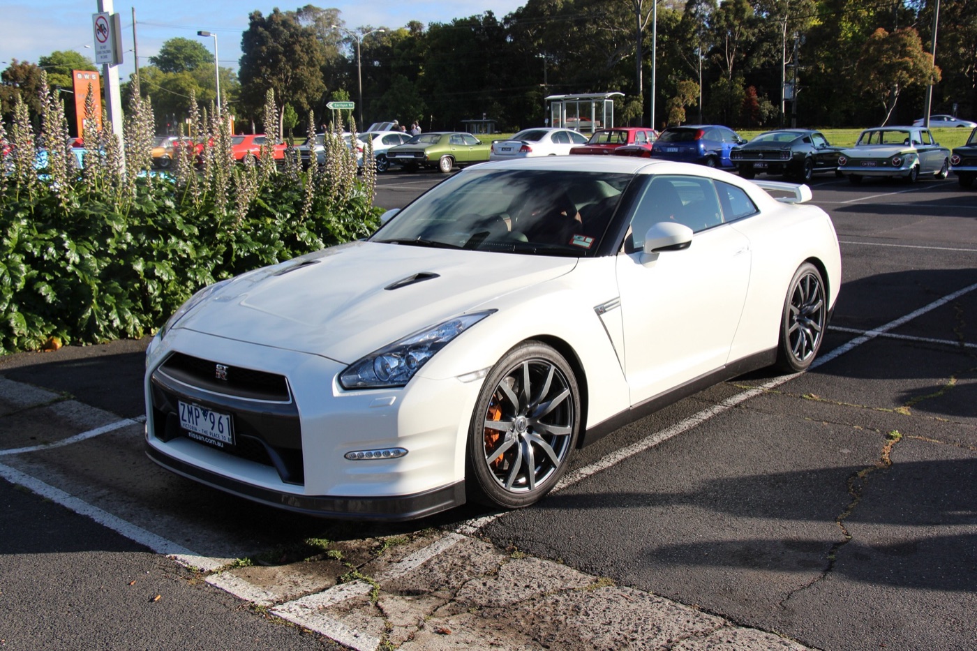 White Nissan GT-R of the 2011 to 2016 shape with ten-spoke grey wheels and orange brake calipers, parked in a car park with older cars behind