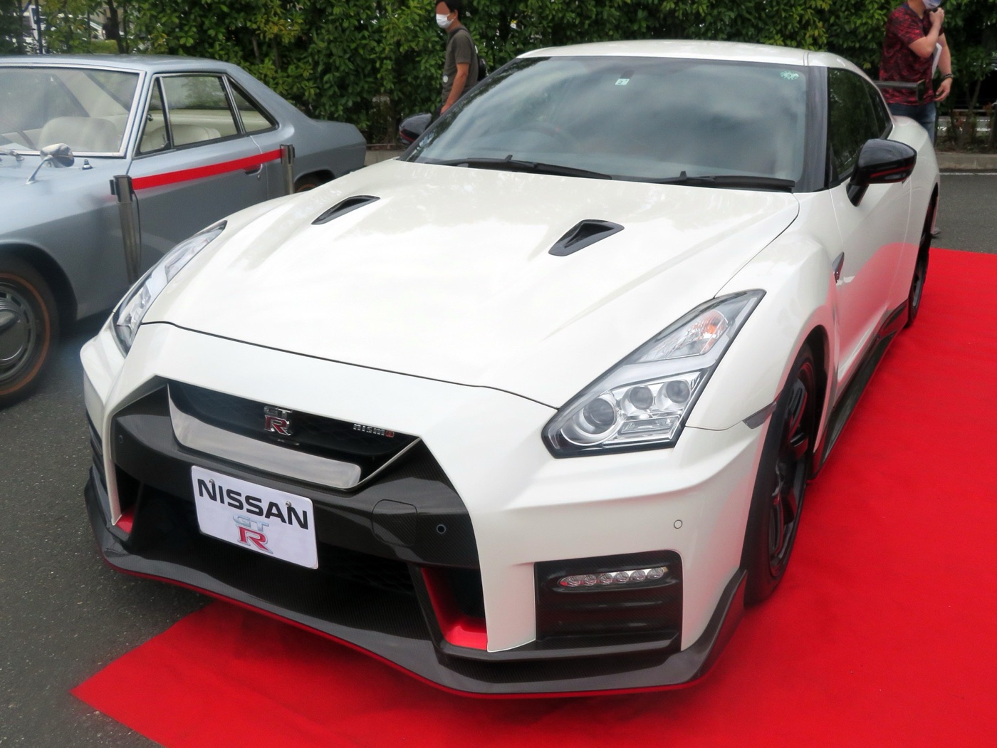 White Nissan GT-R Nismo of the 2017 shape, front view on a red carpet, with carbon-fibre front splitter and red trim, an older Skyline beside it