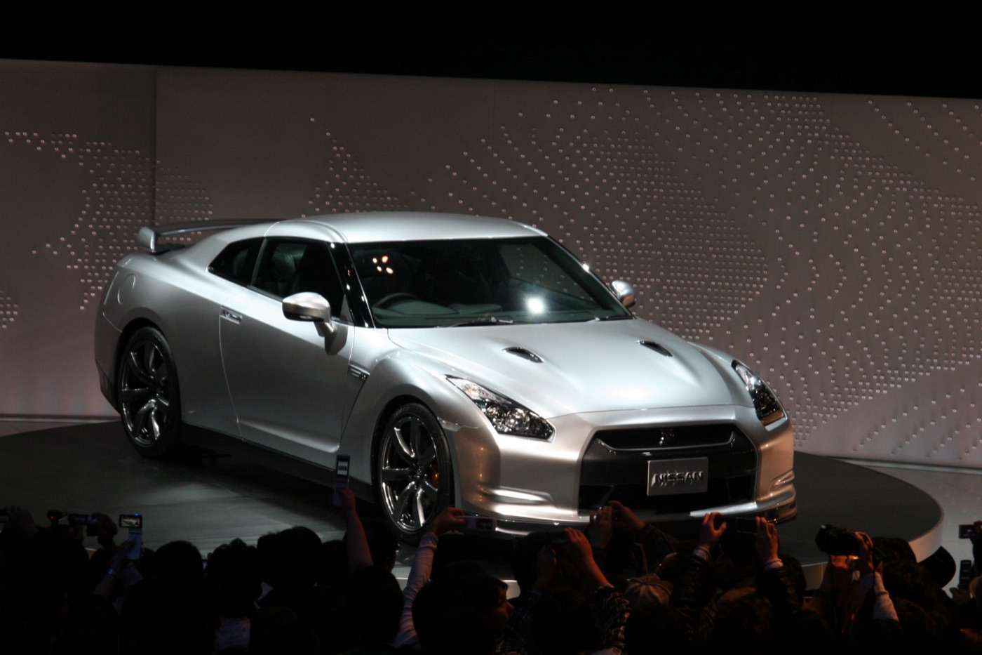 Silver Nissan GT-R on a turntable under stage lights, with a crowd holding up phones and cameras in the foreground