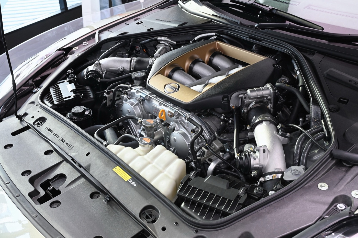 Engine bay of a 2025 Nissan GT-R T-spec with a bronze-coloured cover over the VR38DETT V6 and a carbon-fibre panel marked carbon-fiber composite