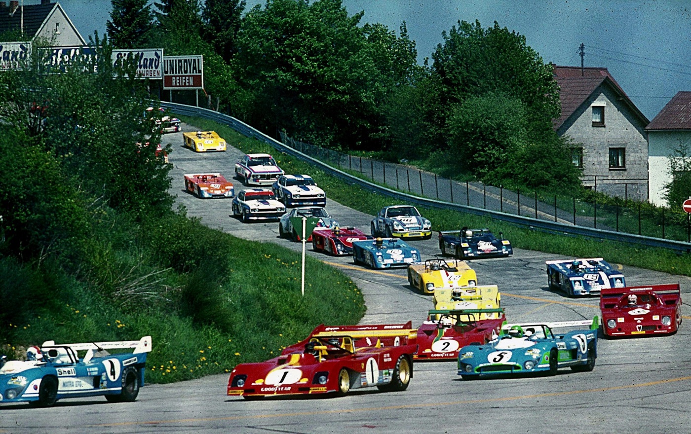 A pack of sports-racing cars led by red Ferrari 312 PBs and blue Matras on the introduction lap, rounding a bend past houses and trees
