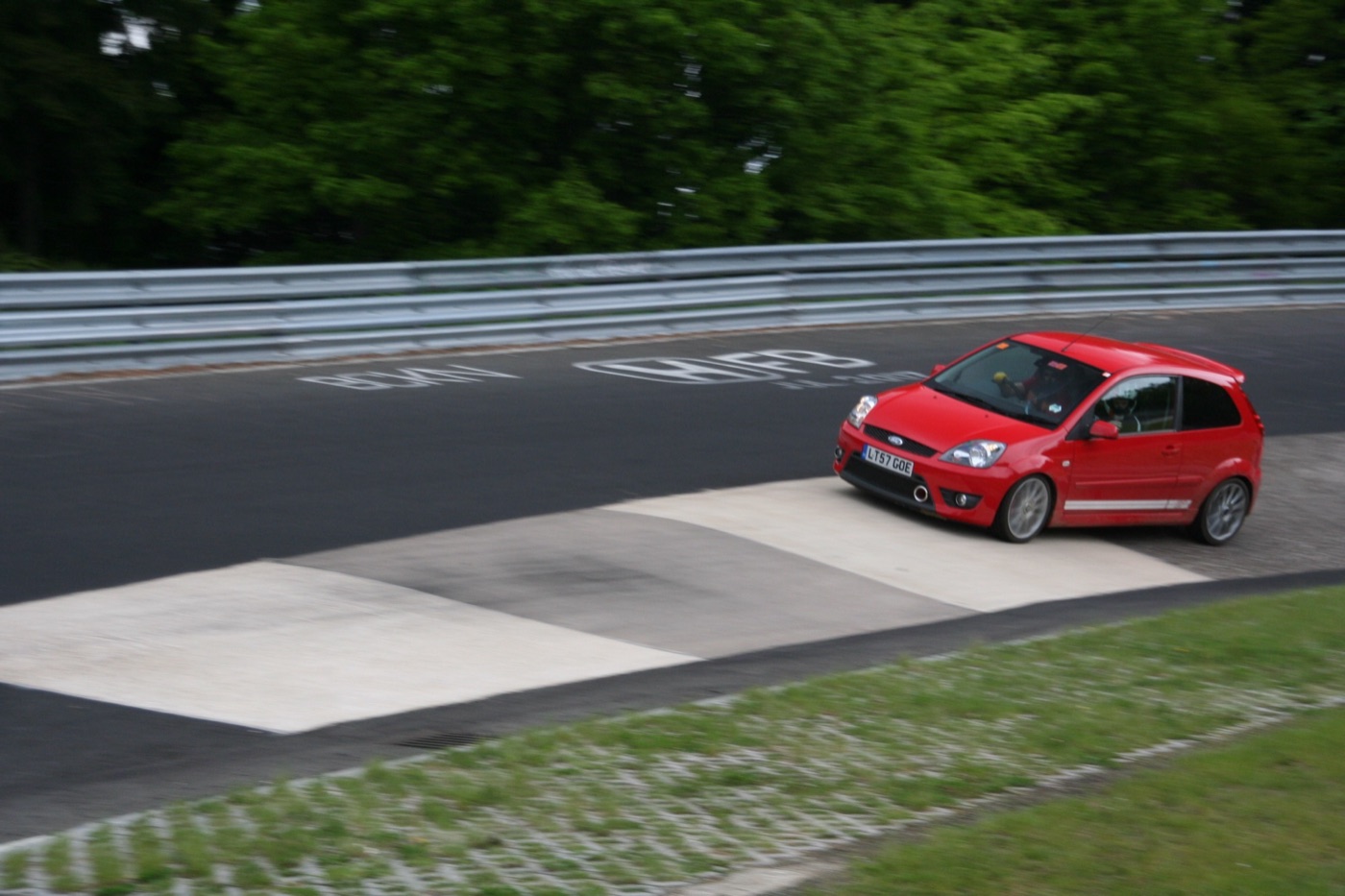 A red Ford Fiesta ST hatchback leaving the concrete banking of the Karussell on the Nordschleife, trees behind the barrier