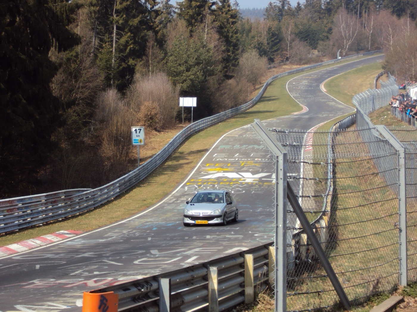 A silver Peugeot hatchback driving through the Pflanzgarten section of the Nordschleife between Armco barriers, spectators behind a fence on the right