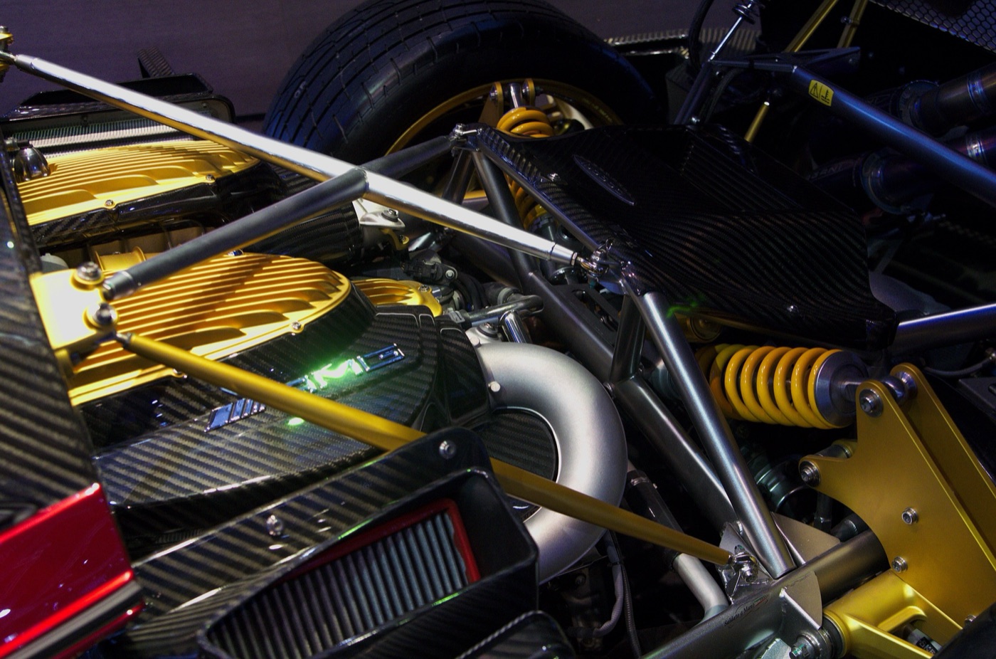 Engine bay of a Pagani Huayra showing gold-finished AMG V12 cam covers, carbon intake parts, a turbo pipe and yellow coil springs