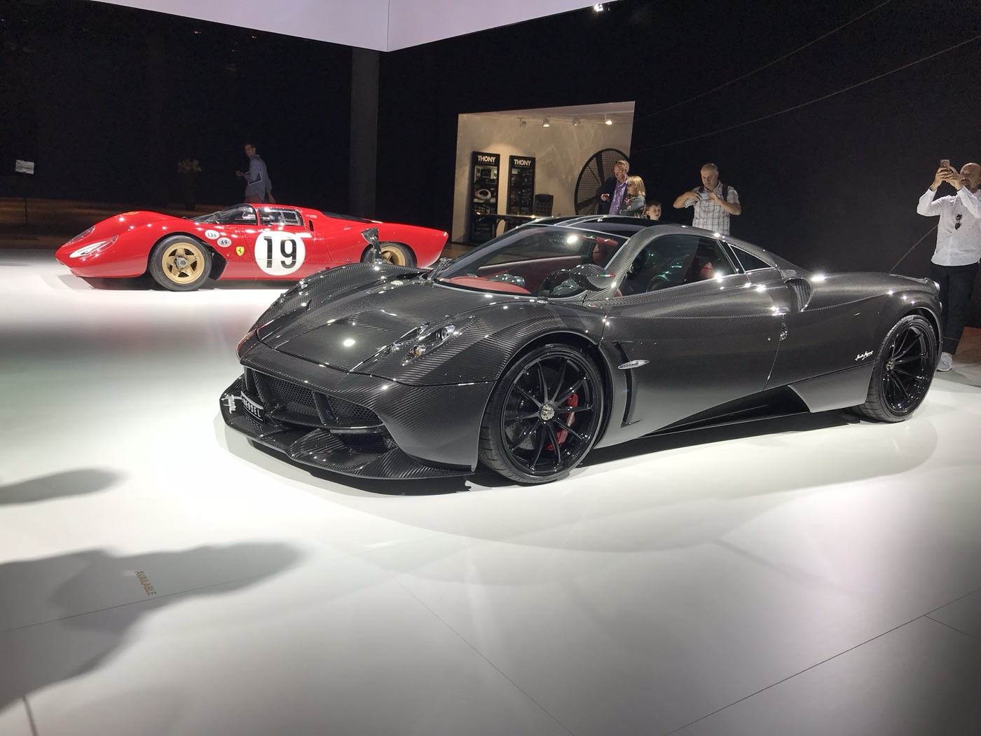 Pagani Huayra coupé in exposed carbon fibre with black wheels and red brake callipers on a white stand, a red Ferrari sports-racer behind