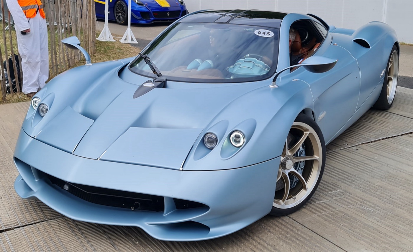 Pale blue Pagani Huayra Codalunga with twin round headlamps and gold wheels, driving slowly through the Goodwood paddock