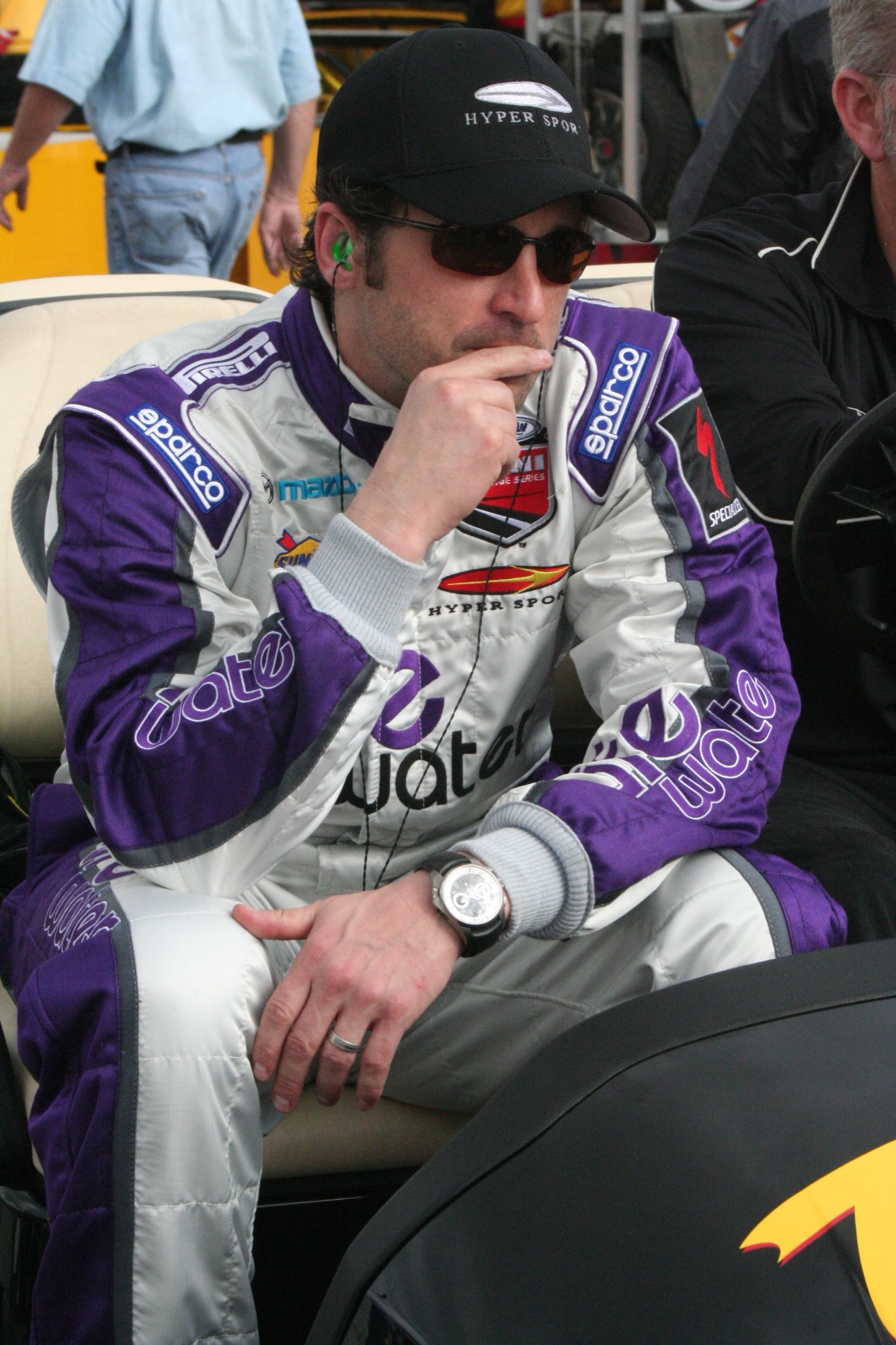 Patrick Dempsey in a white and purple Hyper Sport race suit and cap, seated in the Daytona paddock