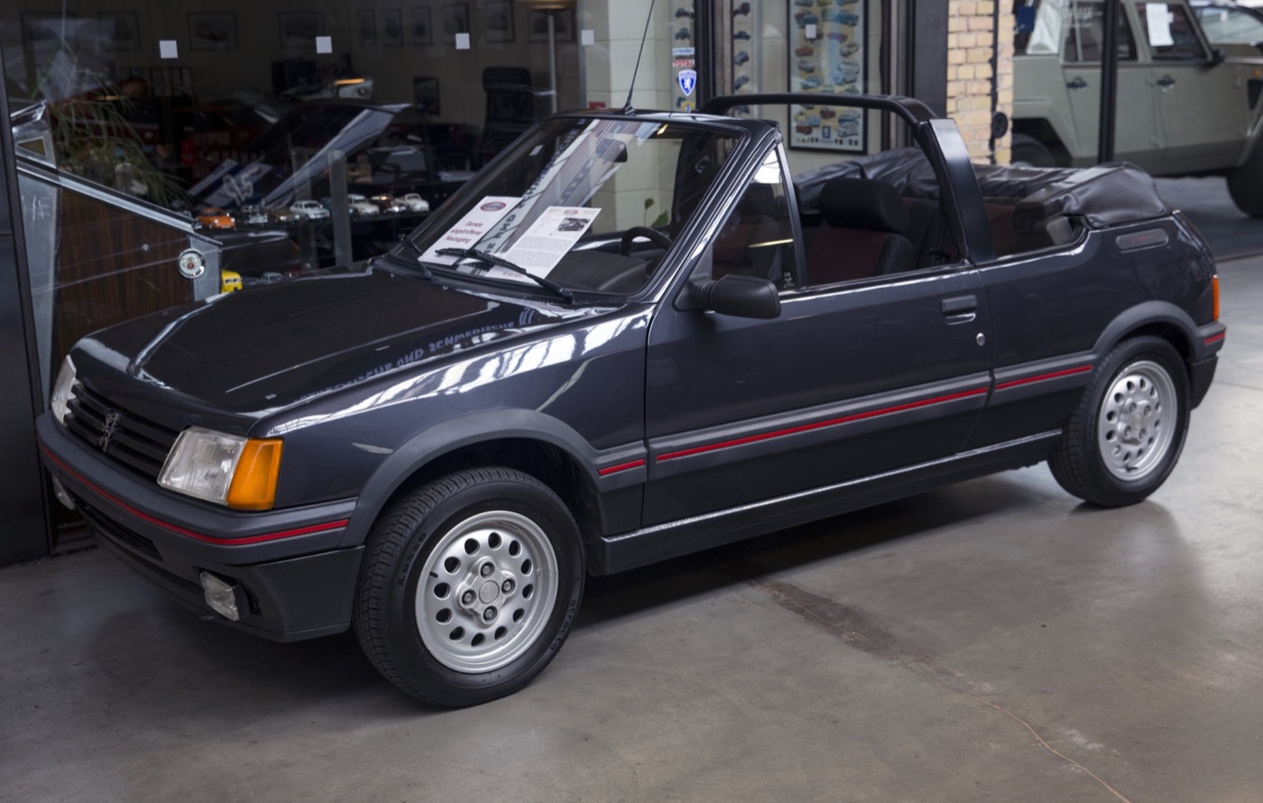 Dark grey Peugeot 205 CTI cabriolet with its roof folded behind a black roll bar, on show inside a classic car dealership