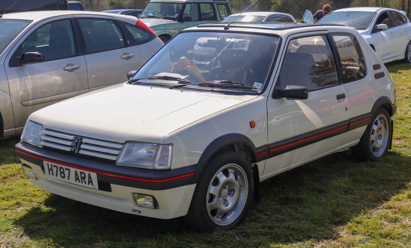 White Peugeot 205 GTi 1.9 with nine-hole alloy wheels and red pinstripes, parked on grass among modern cars