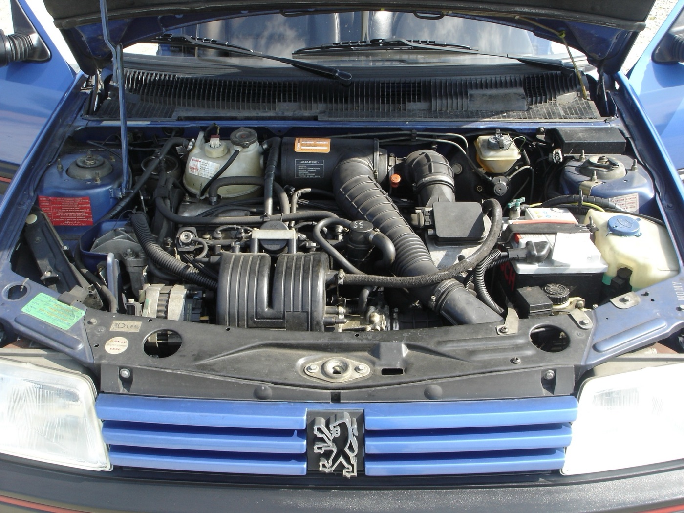 Engine bay of a blue Peugeot 205 GTi 1.9, with the black air intake trunking running across the transverse four-cylinder engine