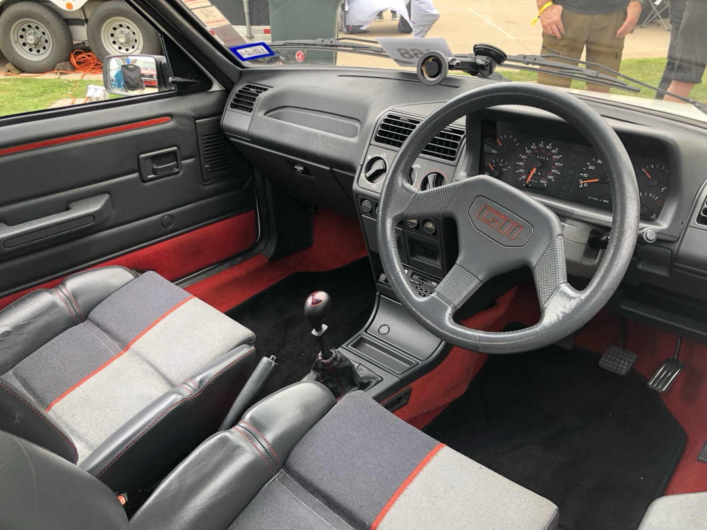 Right-hand-drive Peugeot 205 GTi cabin with a grey three-spoke GTI steering wheel, half-leather seats with red stitching and red carpet