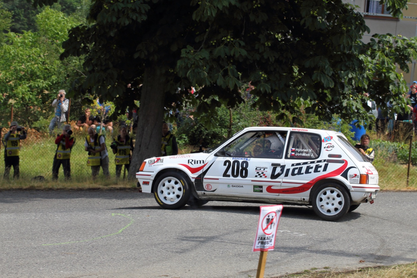 White Peugeot 205 GTi 1.9 rally car with number 208 cornering on tarmac, photographers in yellow vests behind