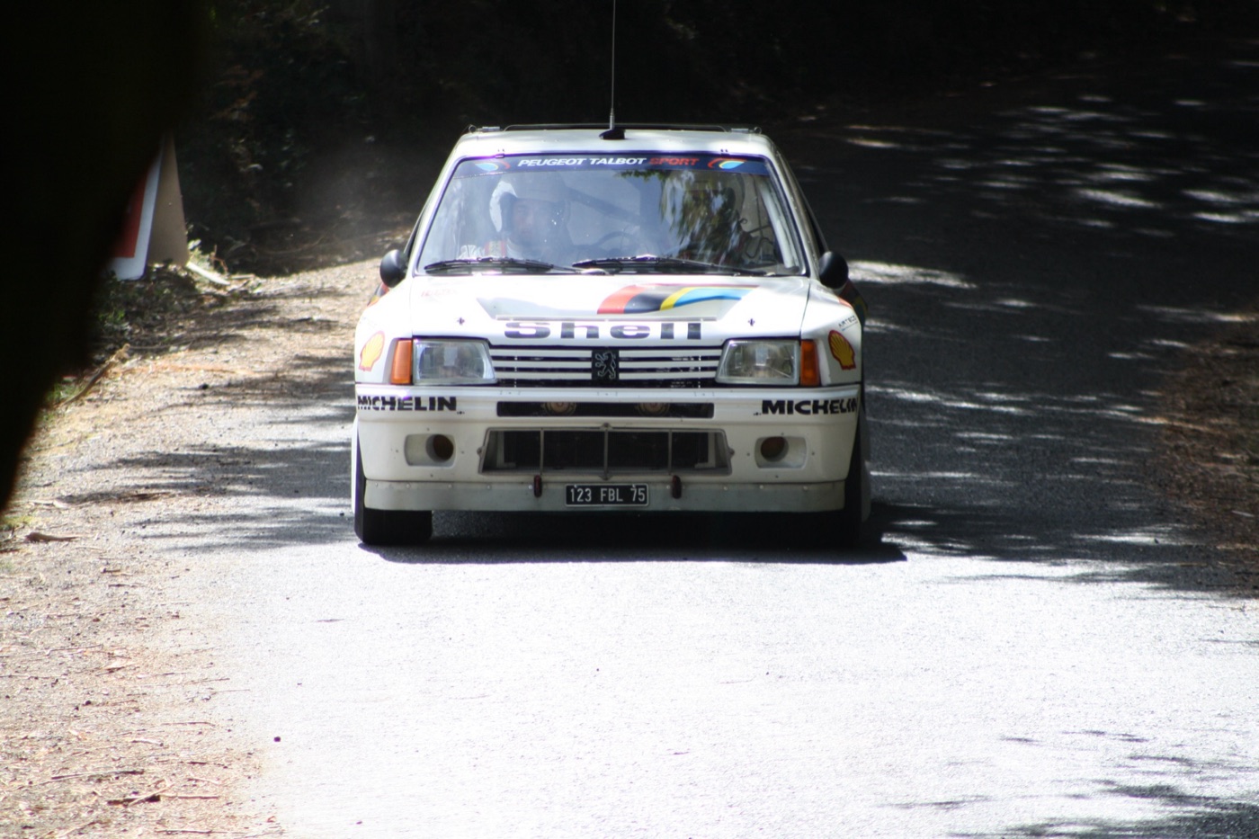 White Peugeot 205 Turbo 16 in Shell and Michelin colours driving towards the camera on a shaded tarmac road