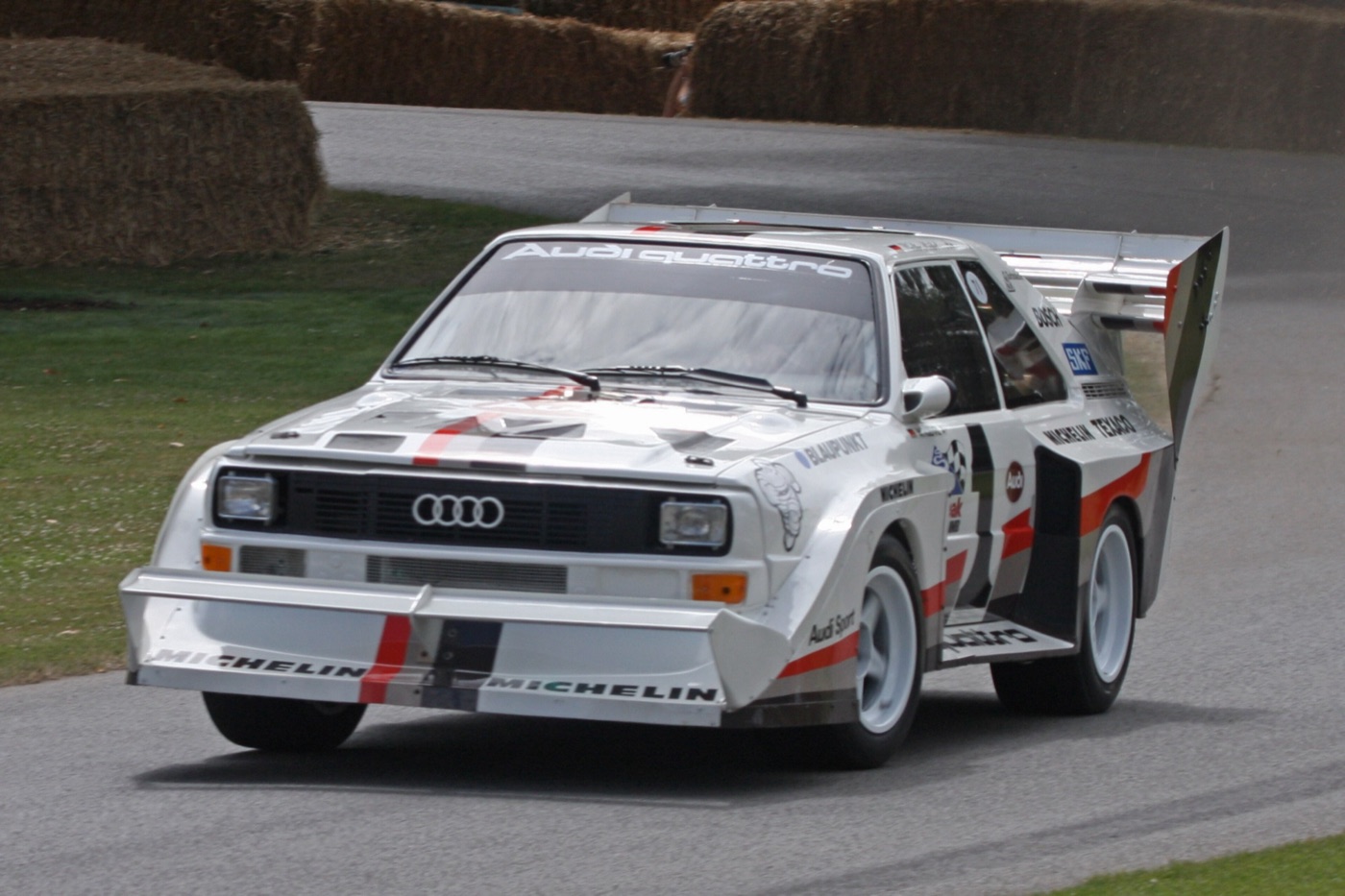 White Audi Sport Quattro S1 with a huge rear wing and front splitter, Michelin and Audi Sport decals, driving past straw bales