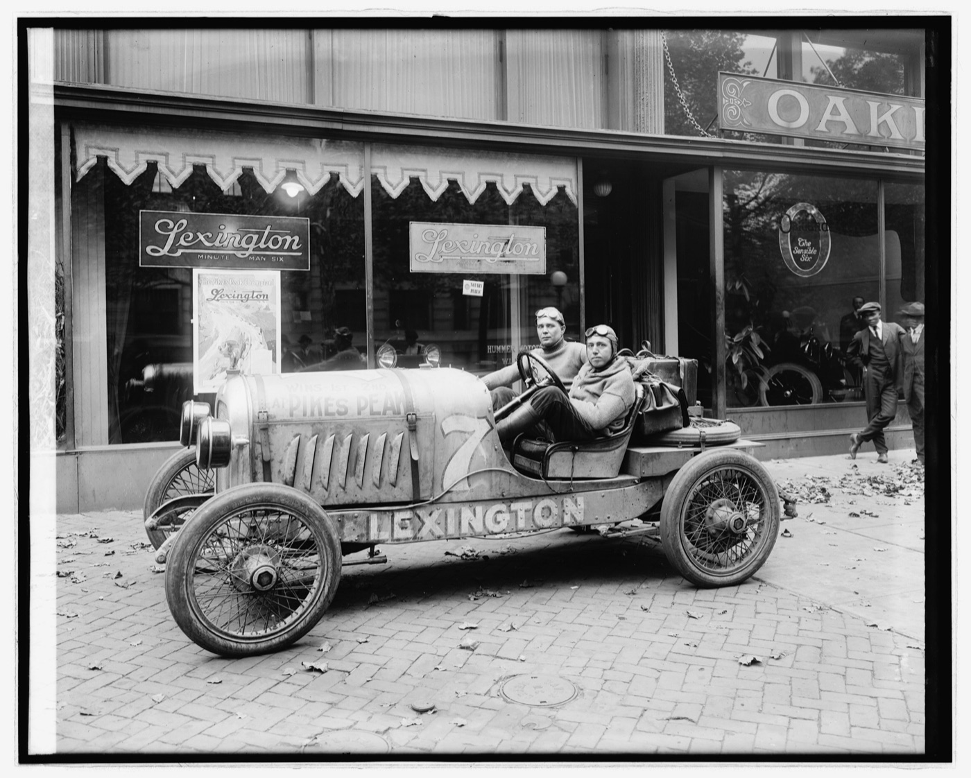 Black and white photograph of a Lexington racing car with Pikes Peak painted on its bonnet and two men aboard, parked outside a Lexington showroom
