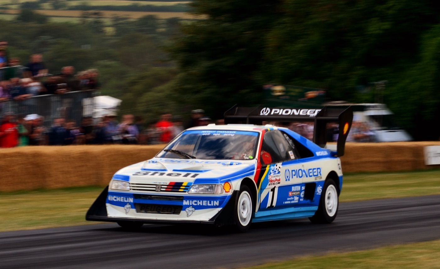 White and blue Peugeot 405 T16 Pikes Peak with a tall rear wing and Pioneer and Michelin decals, driving past a crowd behind straw bales