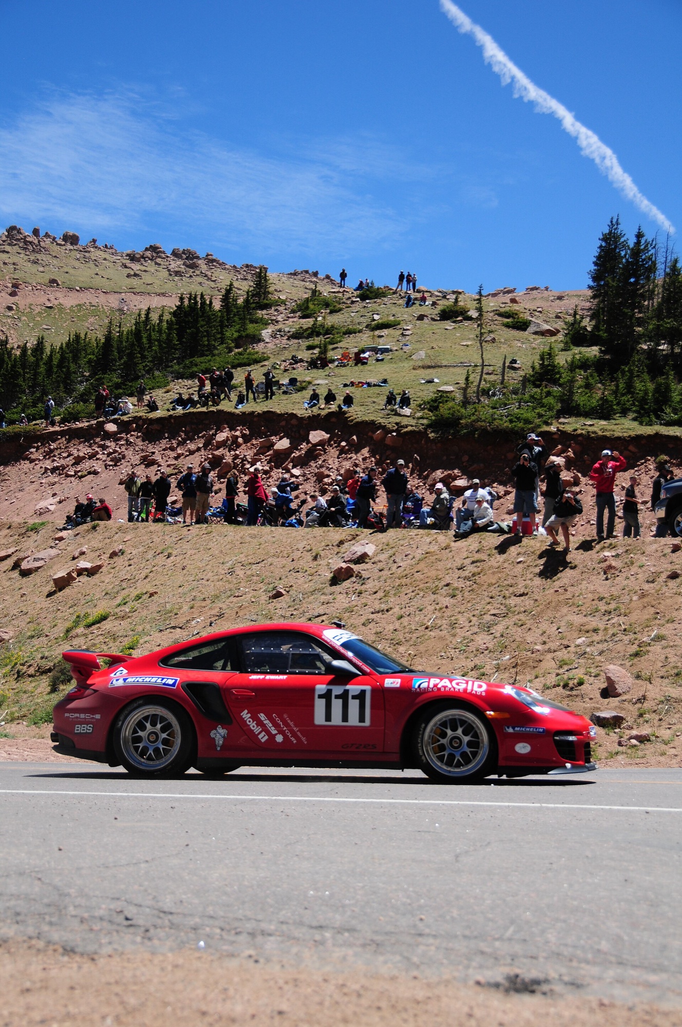 Red Porsche 911 GT2 RS with race number 111 passing a bank of spectators sitting on the rocky hillside above the road