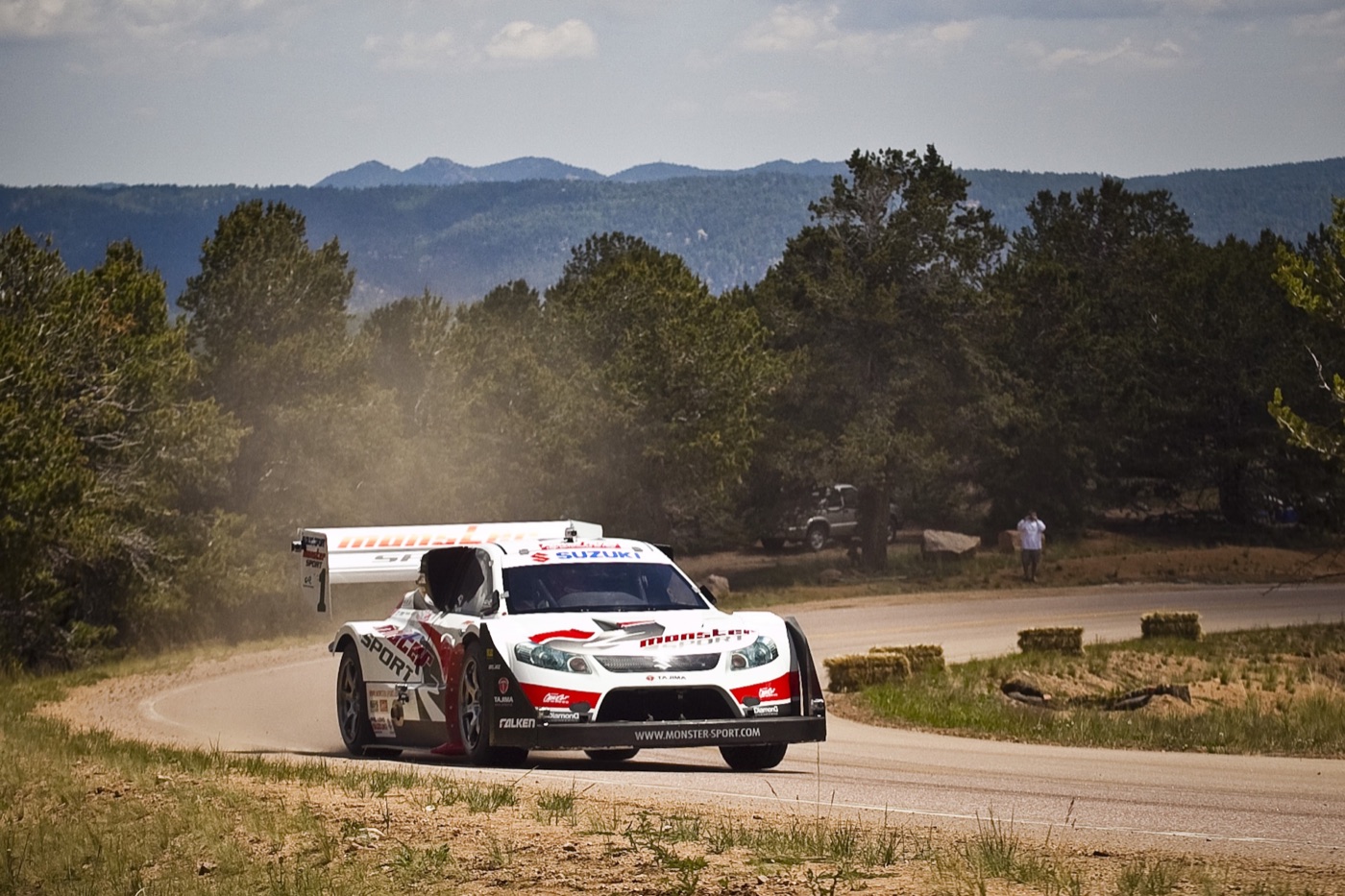 White and red Suzuki SX4 Hill Climb Special with a high rear wing on a forested bend of the Pikes Peak course, with dust behind it