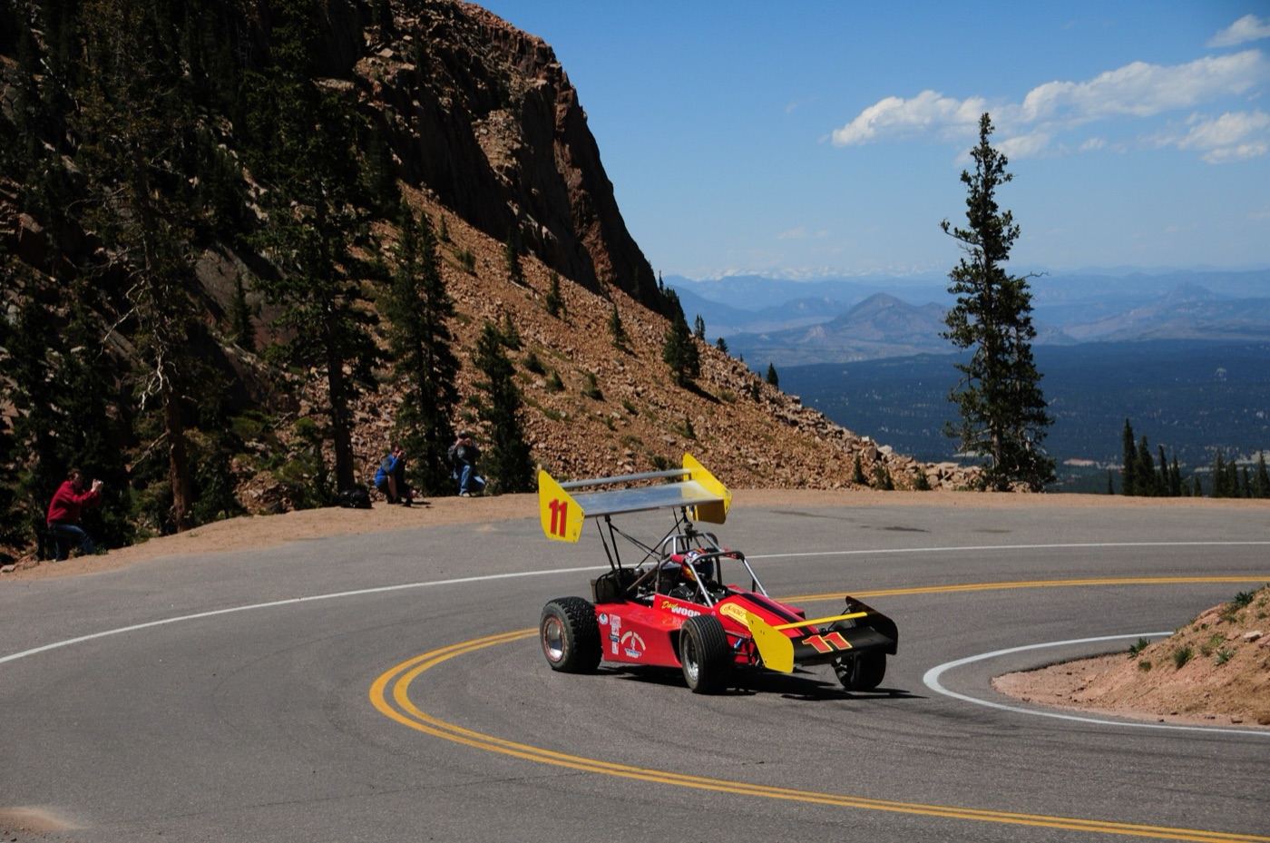 Red and yellow Wells Coyote open-wheel racer with a high rear wing rounding a paved hairpin on Pikes Peak, with spectators on the slope and the plains far below