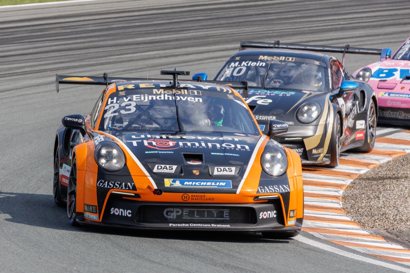 Orange and black Porsche 911 GT3 Cup number 23 leading a black and gold 911 GT3 Cup number 10 through a corner at Zandvoort